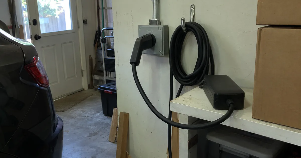 A portable EV charging unit plugged into a 240-volt receptacle in a gray metal box on a garage wall, with conduit above and the cable coiled on a hook