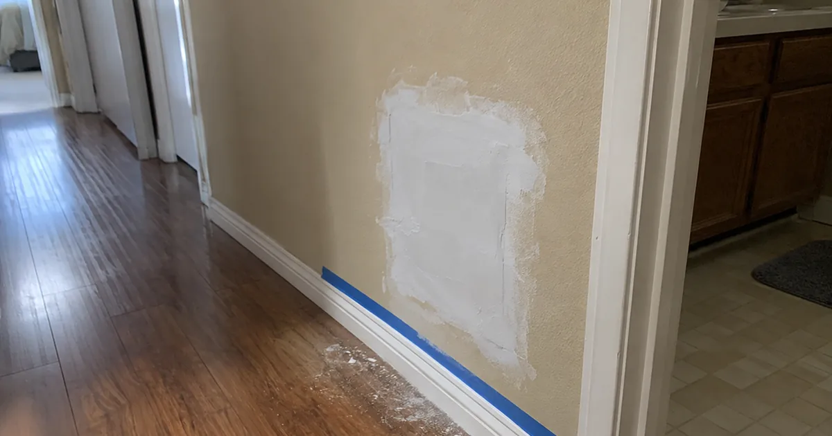 A freshly closed, unpainted drywall patch low on a hallway wall beside a bathroom door, with blue painter's tape on the baseboard and compound dust on the floor