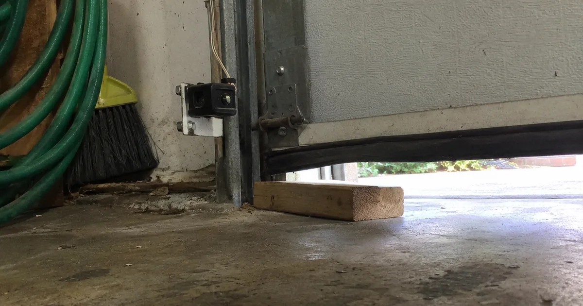 Low view across a garage floor of a photo-eye safety sensor on the door track and a short block of 2x4 lumber lying under the partly lowered garage door